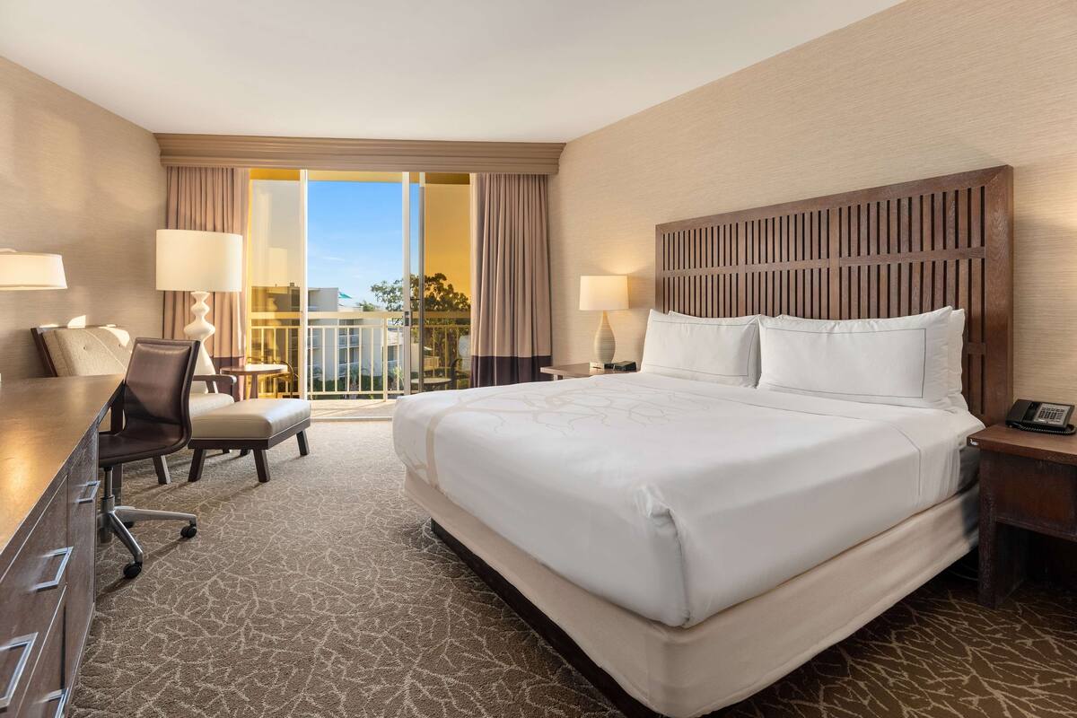 A hotel room with a large bed, a desk, a chair, and a balcony with curtains.