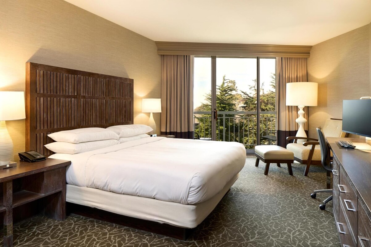 A hotel room with a large bed, a desk with a computer, a chair, a telephone, and a view of trees through the window.