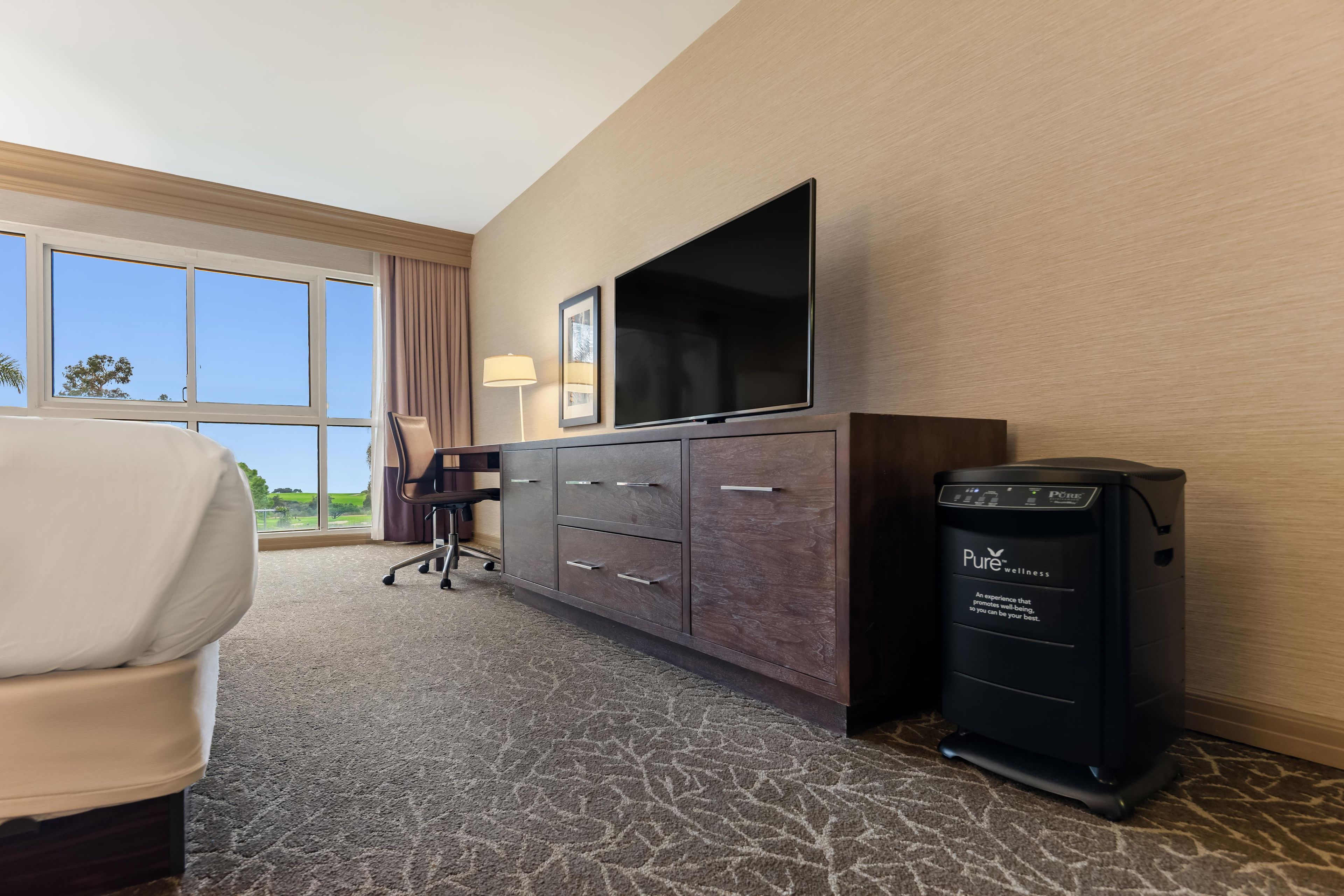 A hotel room with a large window, a flat-screen TV, a desk with a chair, a minibar, and a carpeted floor.