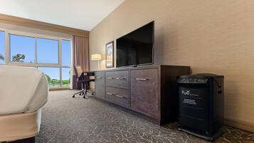 A hotel room with a large window, a flat-screen TV, a desk with a chair, a minibar, and a carpeted floor.