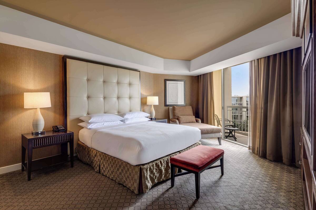 A hotel room with a large bed, a bedside table with a lamp, a chair, and a view of the outdoors.