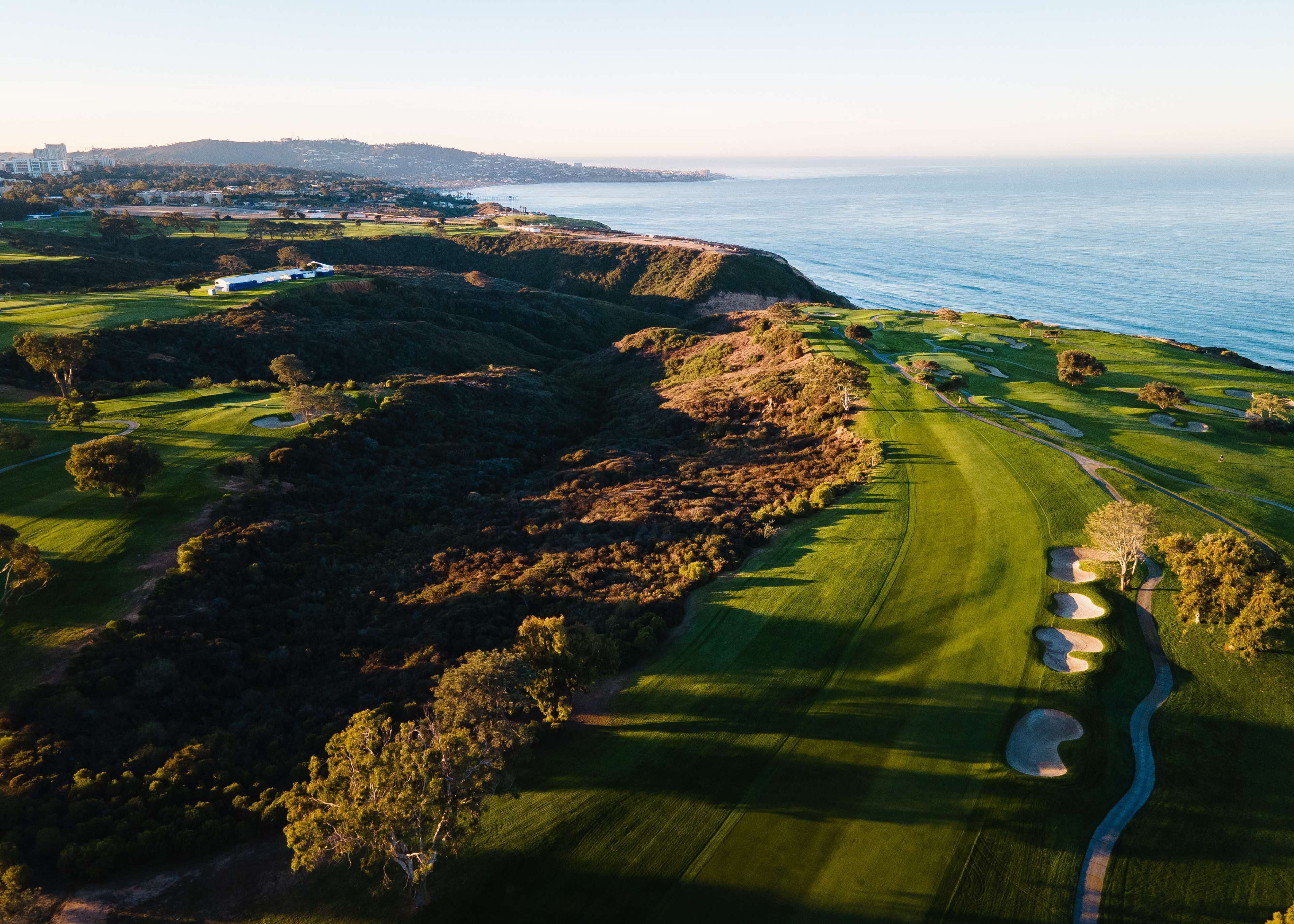 An aerial view of a golf course with a coastal backdrop, featuring well-maintained greens, sand bunkers, and a clear path for play.