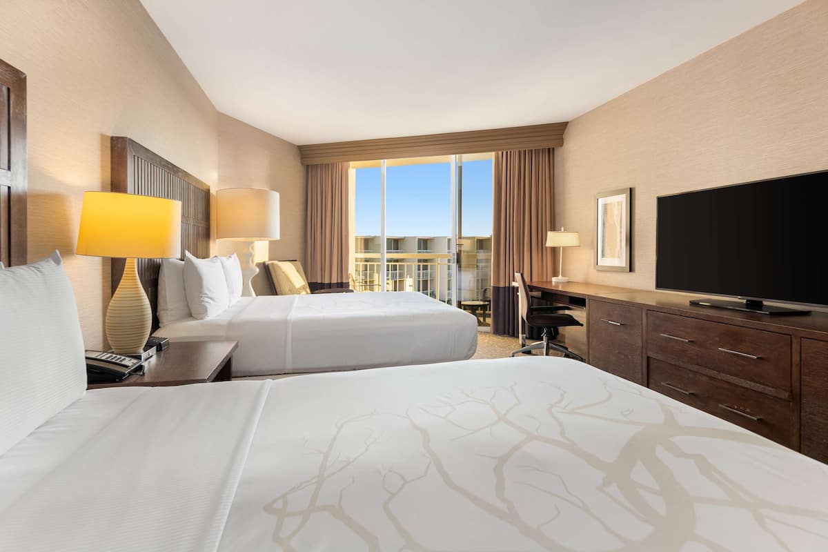 A hotel room with two beds, a desk, a television, and a large window.