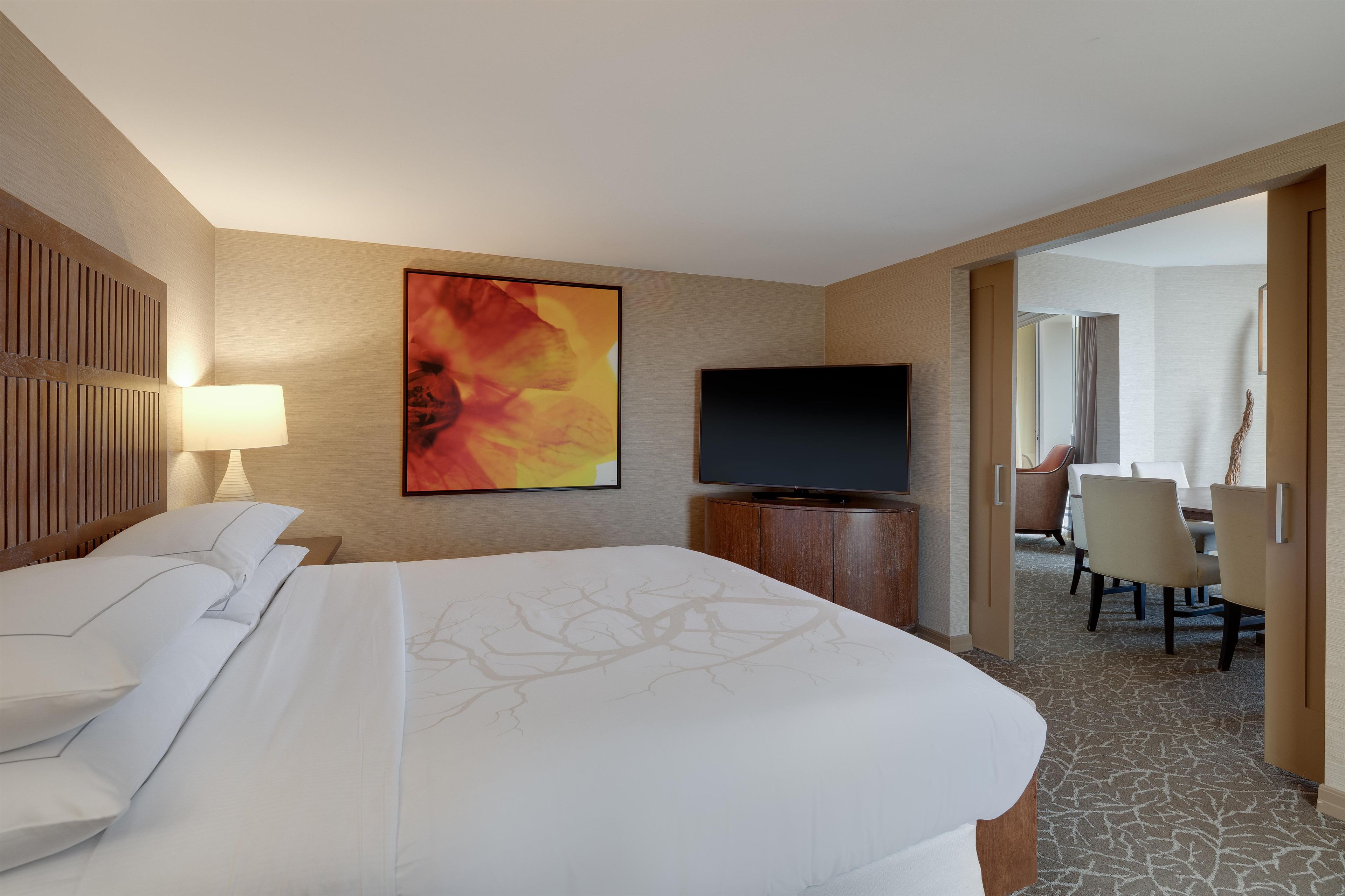 A hotel room with a bed, a television, a painting, and a dining area with chairs and a table.