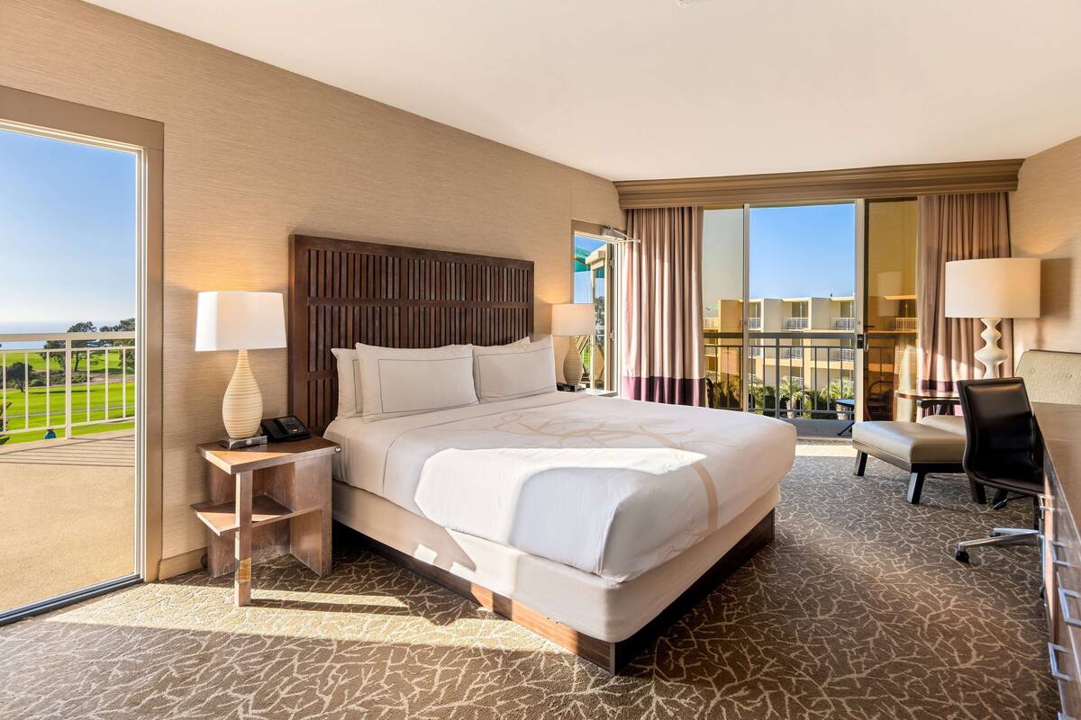 A hotel room with a large bed, a desk, a chair, a lamp, and a view of a golf course.