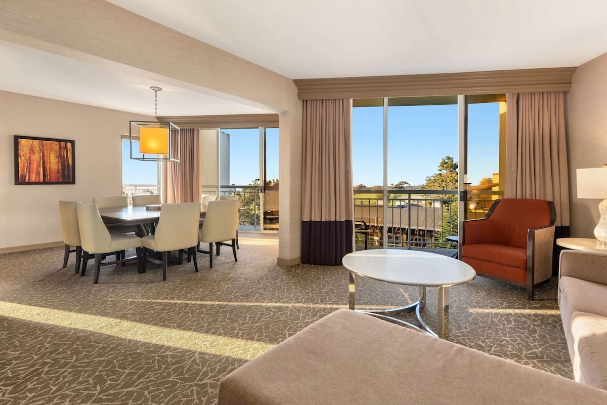 A hotel room with a dining area, a seating area with a table and chairs, and large windows offering a view.