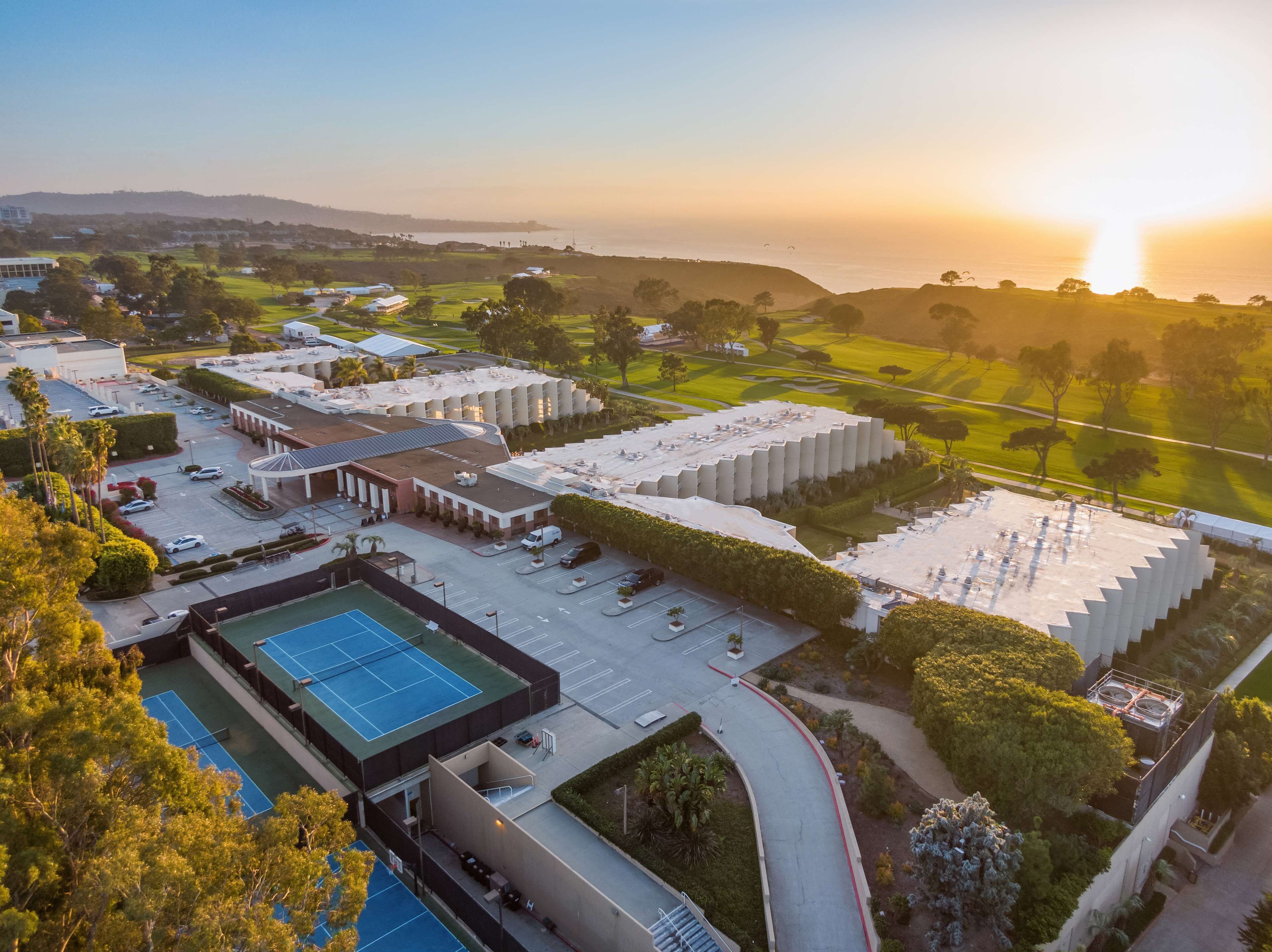 An aerial view of a hotel with multiple buildings, a tennis court, and a golf course in the background.