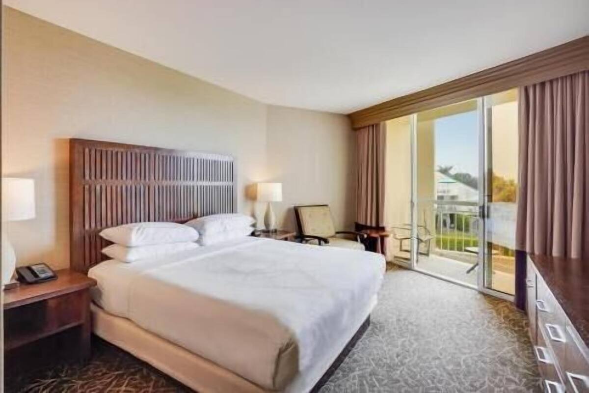 A hotel room with a large bed, a nightstand, a desk with a chair, and a view of the outdoors through a sliding door.