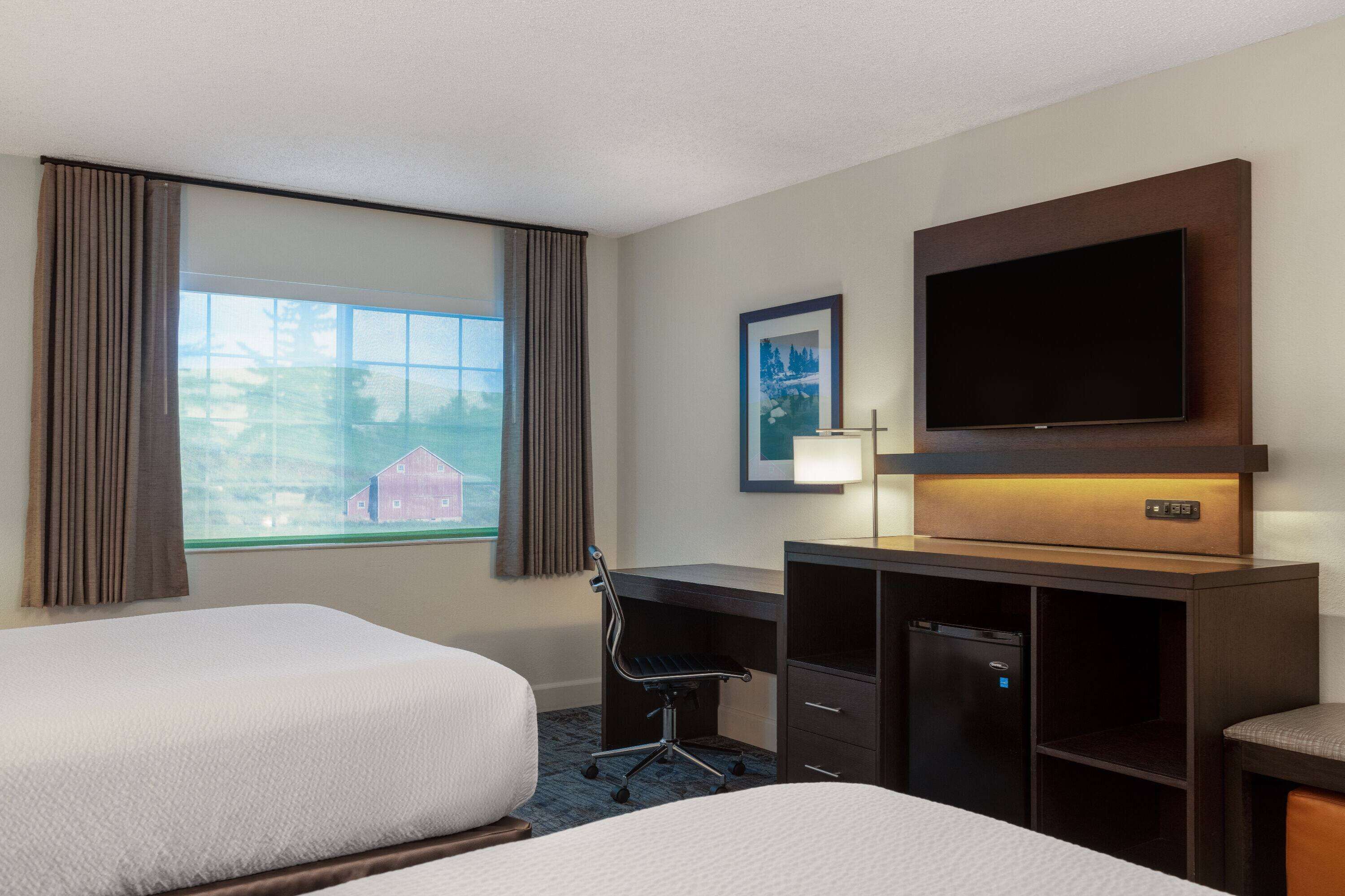 Ein Hotelzimmer mit zwei Betten, einem Schreibtisch mit Computer, einem Fernseher und einem Fenster mit Vorhängen.