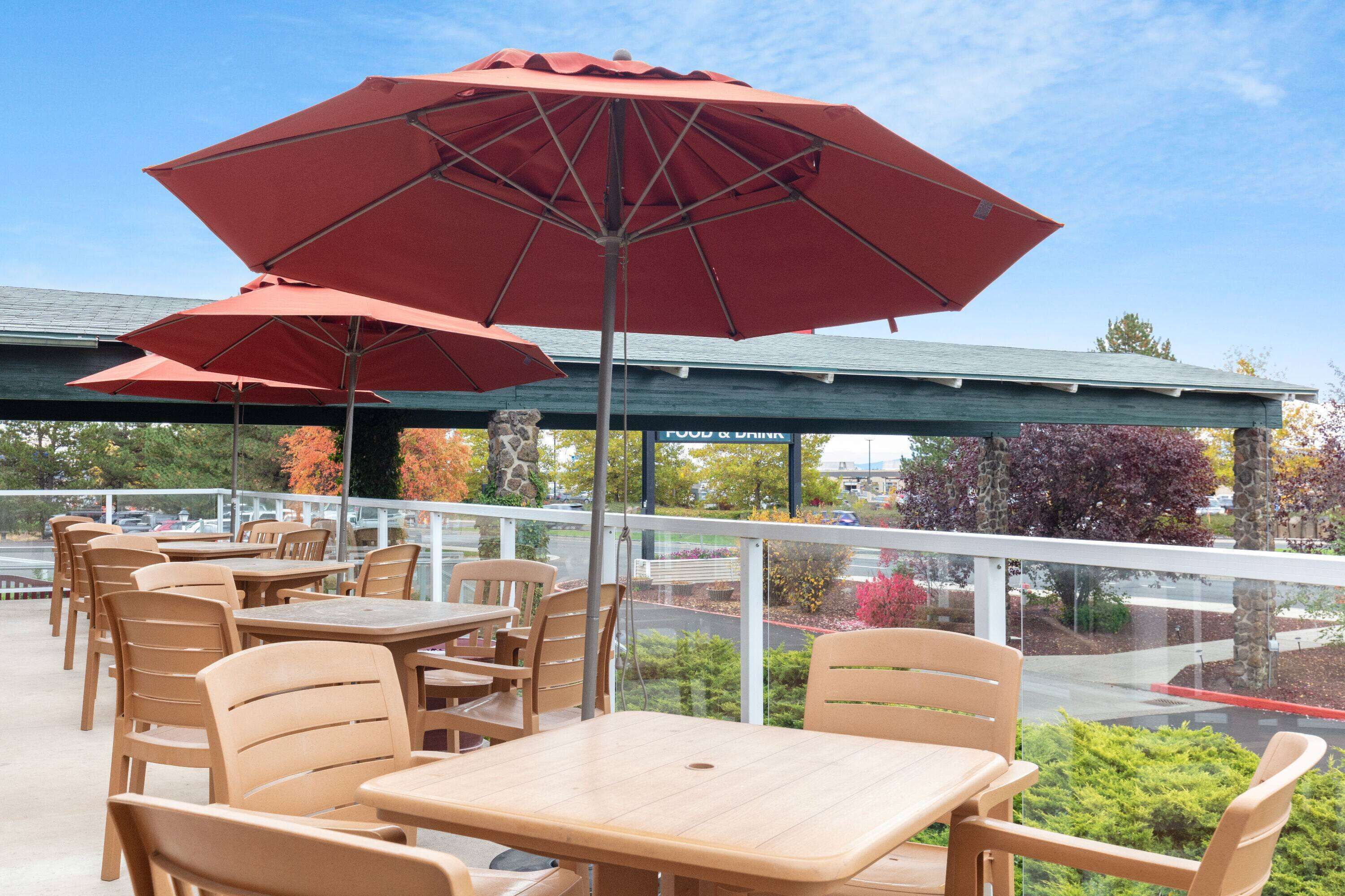 An outdoor patio with wooden tables and chairs, red umbrellas, and a view of the parking lot and trees.