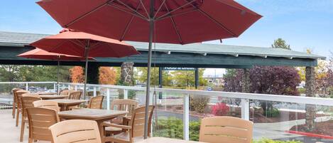 An outdoor patio with wooden tables and chairs, red umbrellas, and a view of the parking lot and trees.