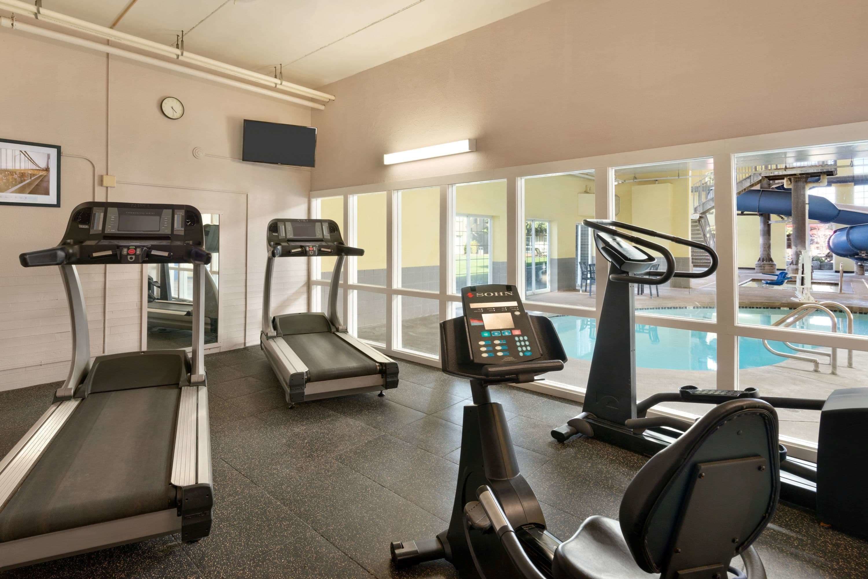 A gym with treadmills, a stationary bike, and a pool area visible through large windows.