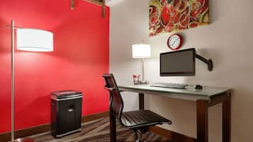 A hotel room with a red accent wall, a desk with a computer, a chair, a trash bin, a floor lamp, and a wall clock.