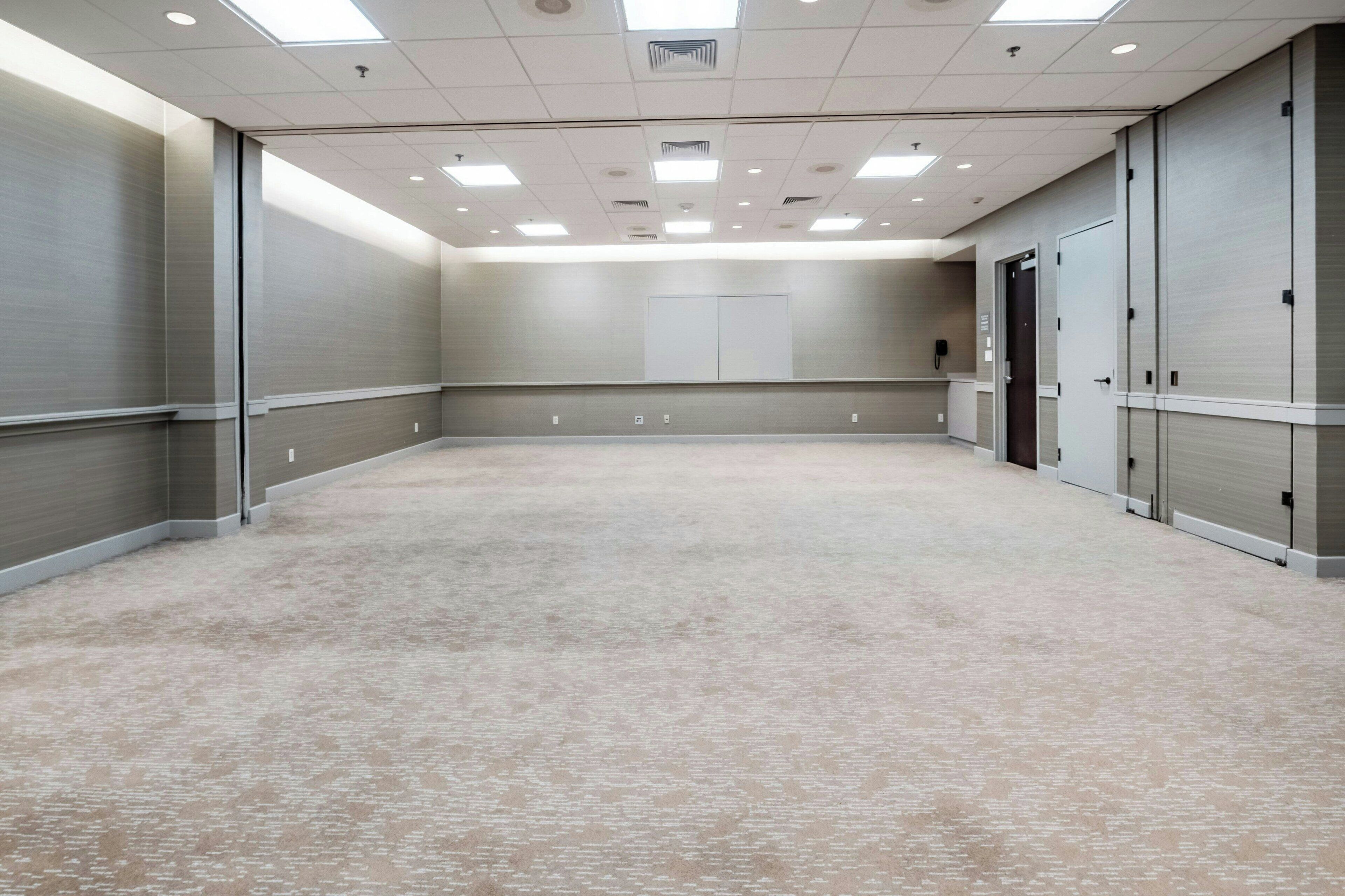 Un salón de conferencias amplio y luminoso, con suelo alfombrado, ventanas grandes y una puerta que da a otra habitación.