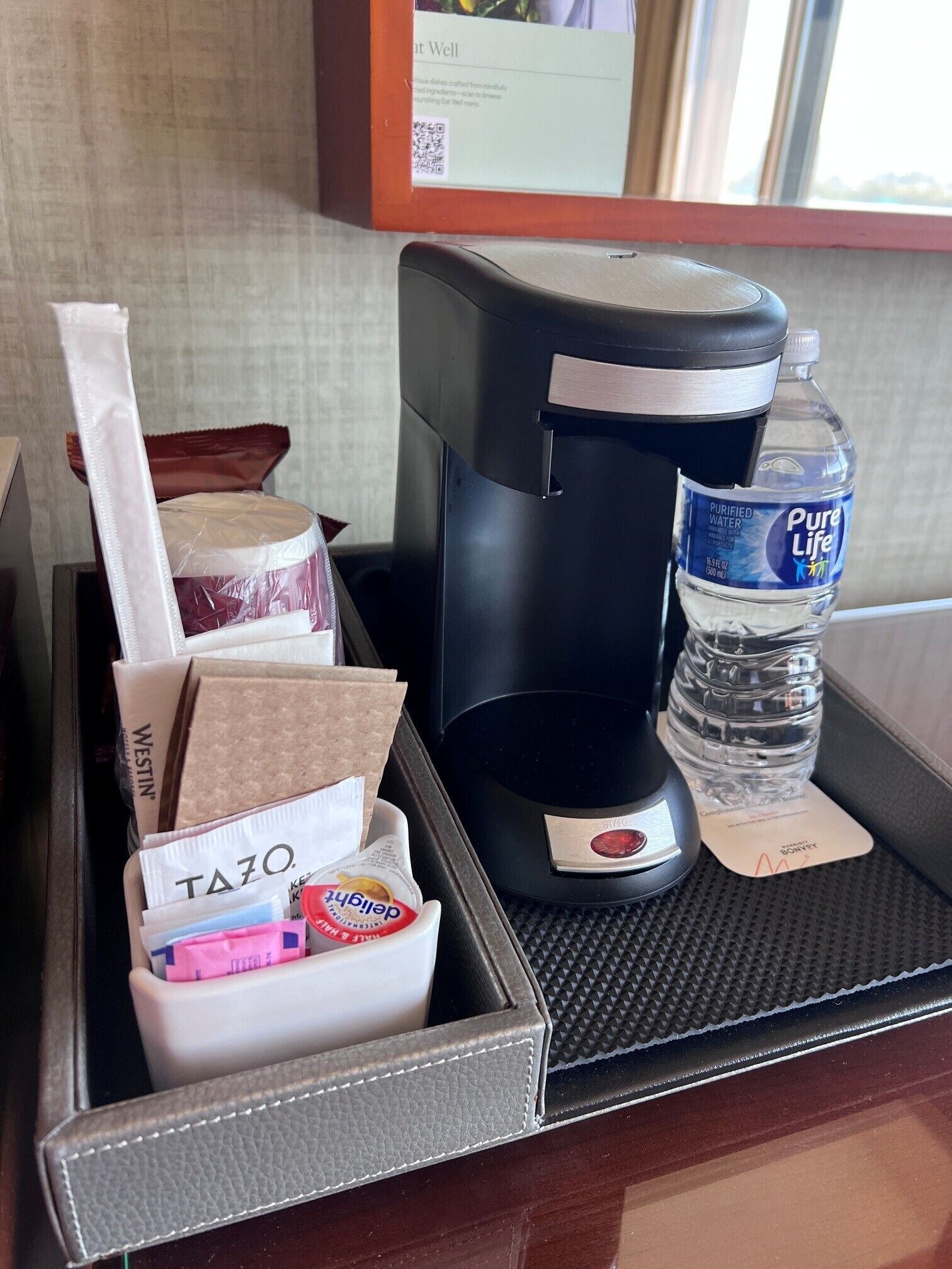 Minibar de una habitación de hotel con una cafetera, una botella de agua y diversos productos alimenticios.