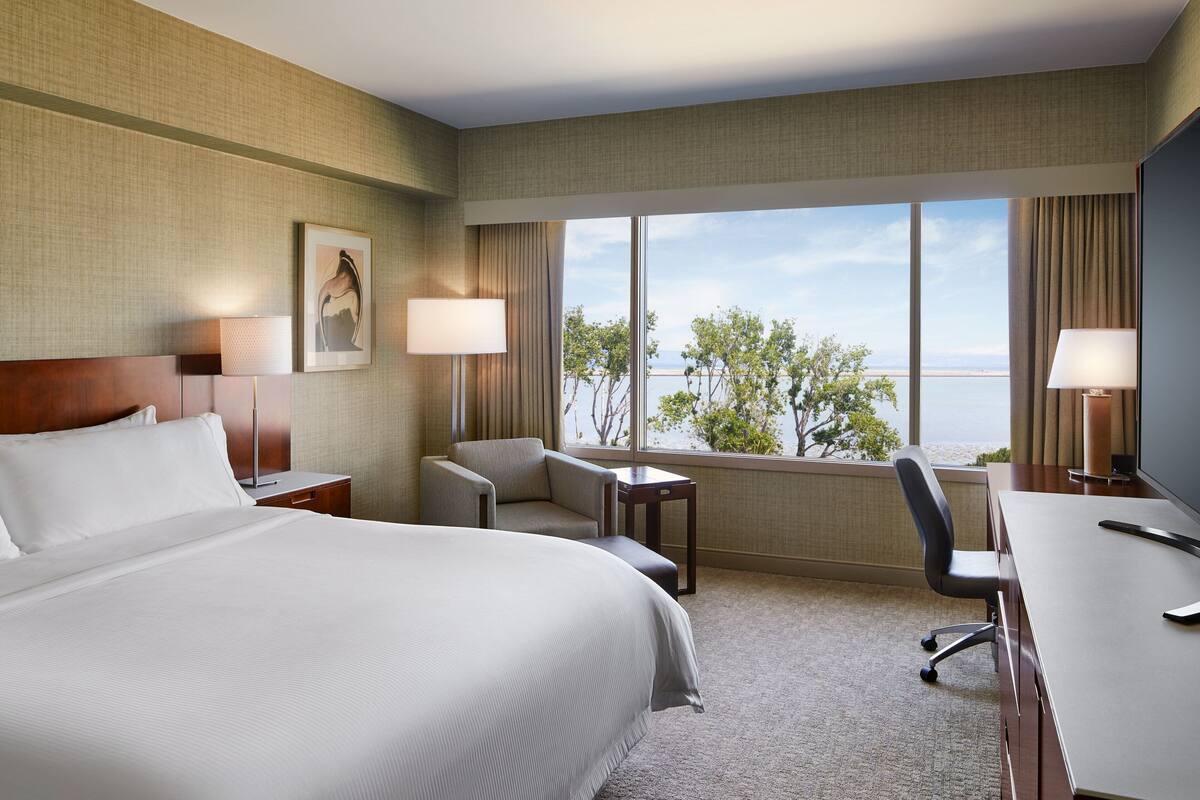 Une chambre d’hôtel avec un grand lit, un bureau, deux chaises, une petite table et une vue sur la mer.
