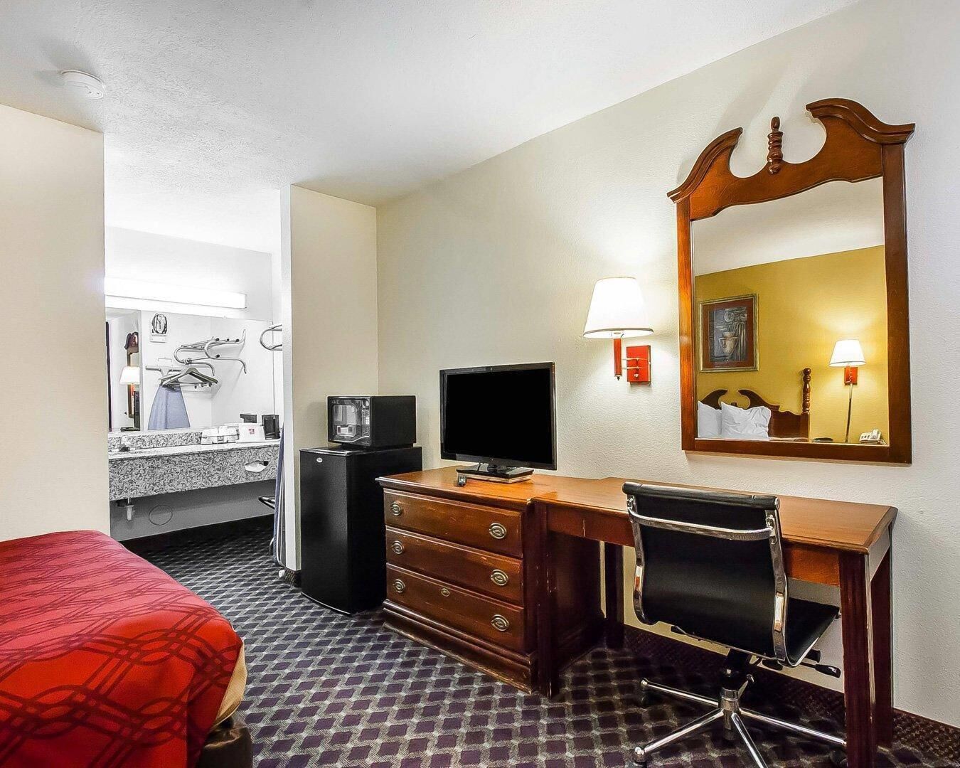 Una camera d'albergo con un letto, una scrivania con un computer, una televisione, uno specchio, una lampada e un bagno sullo sfondo.