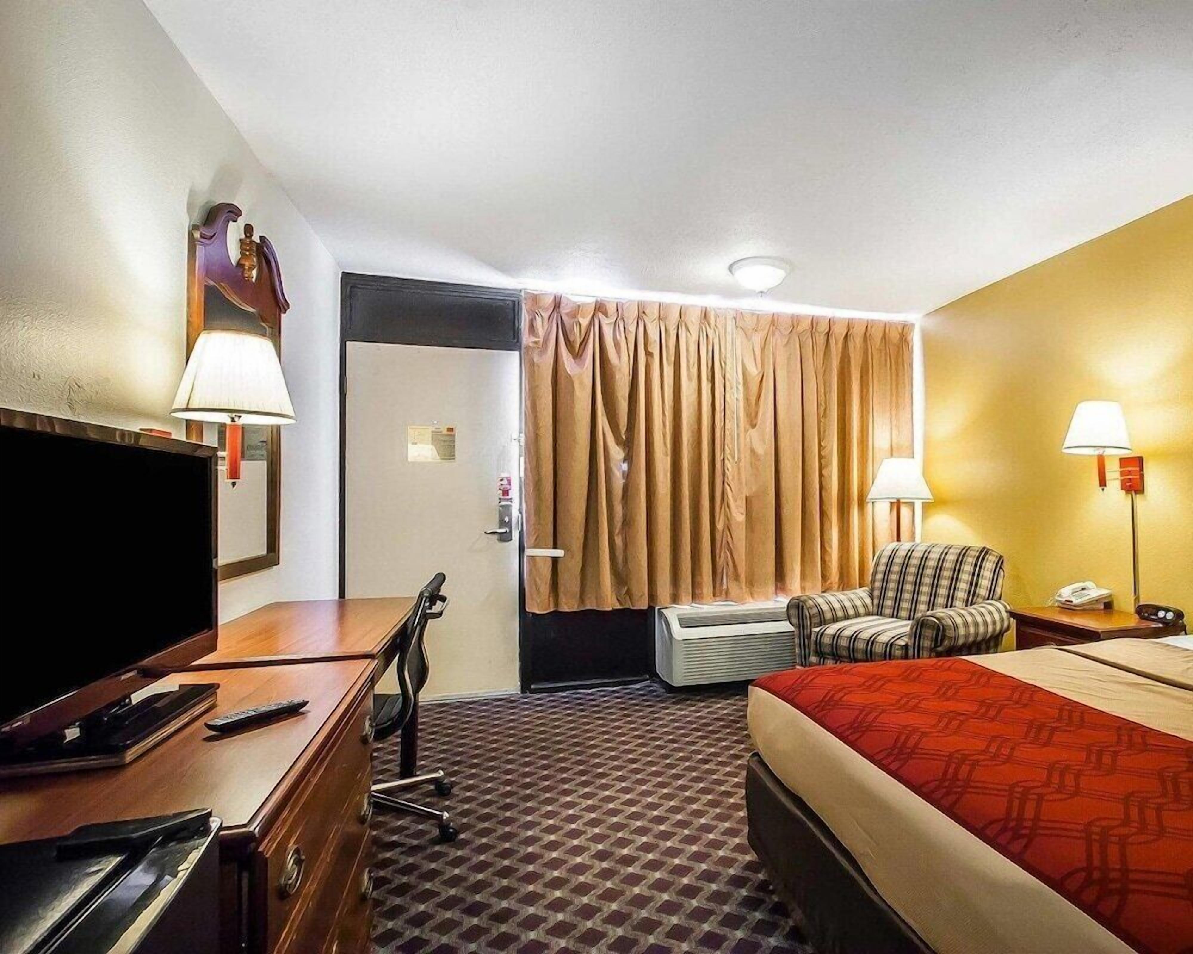 A hotel room with a TV, desk, chair, bed, and lamps.