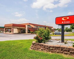 Un hotel con fachada roja y color crema, un estacionamiento y un letrero que indica la entrada.