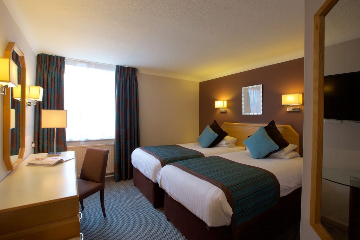 Ein Hotelzimmer mit zwei Betten, einem Schreibtisch und einem Stuhl. Es gibt zwei Nachttische, ein Fenster mit Vorhängen und einen Fernseher an der Wand.