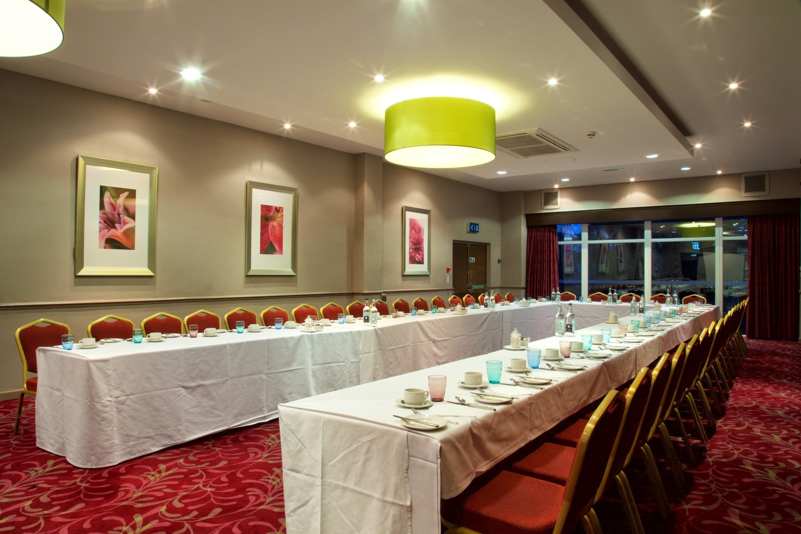 A conference room with a long table set for a meeting, surrounded by chairs, with framed artwork on the walls and a large window with curtains.
