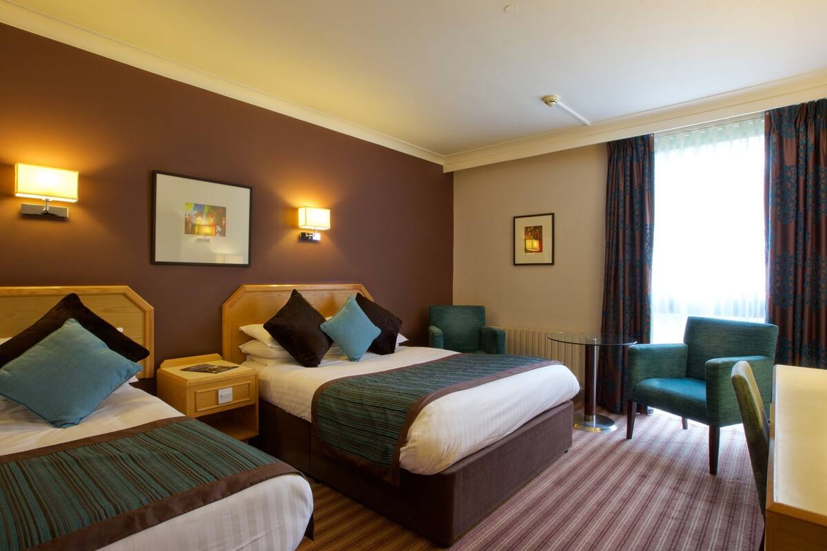 Ein Hotelzimmer mit zwei Betten, einer Sitzecke mit einem Sessel, einem kleinen Tisch und einem Fenster mit Vorhängen.