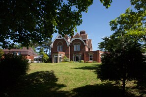 Une grande maison en briques rouges, dotée d’une entrée imposante, et entourée d’une végétation luxuriante et d’arbres.