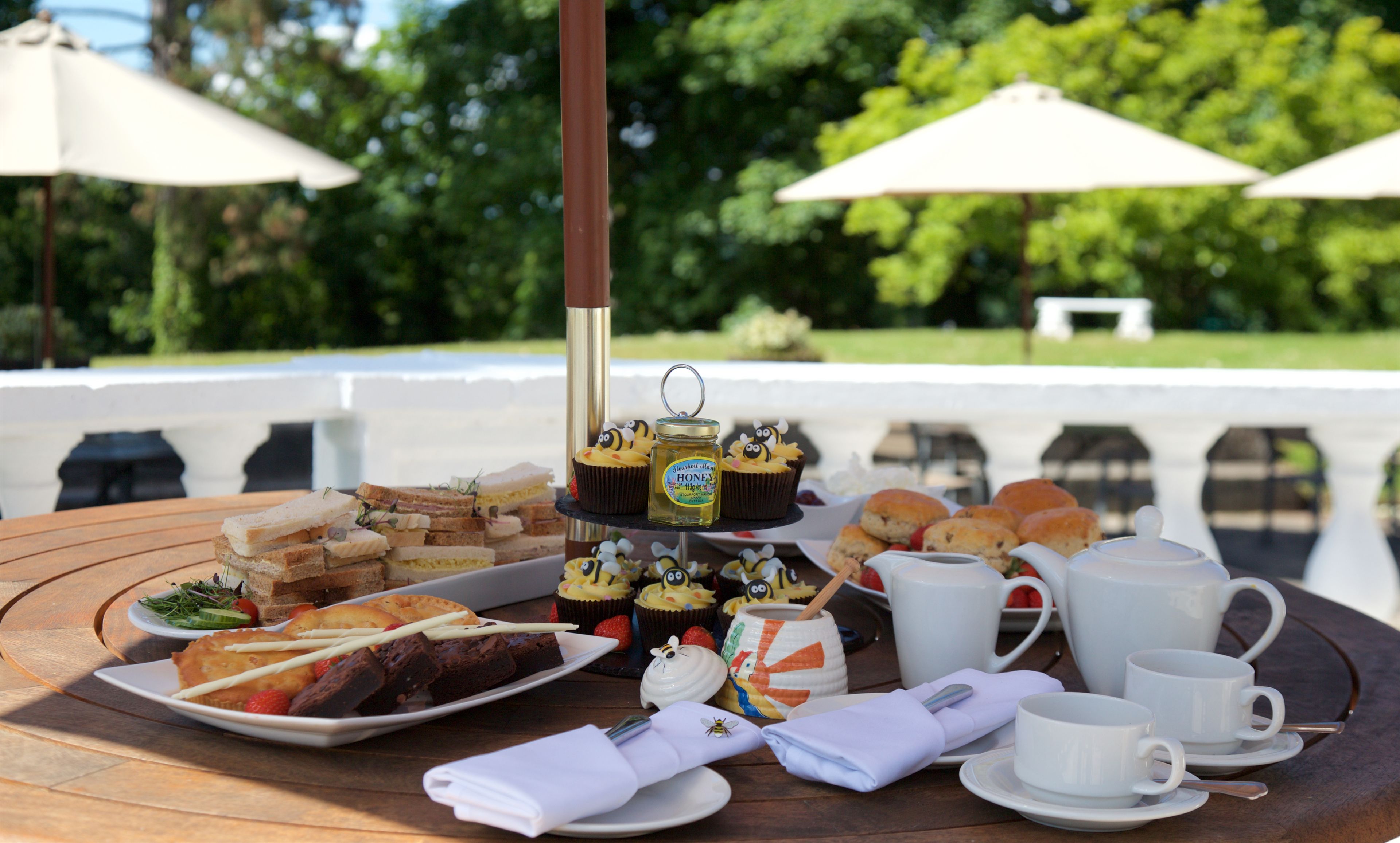A table set outdoors with a variety of food items, including sandwiches, pastries, and a honey jar.
