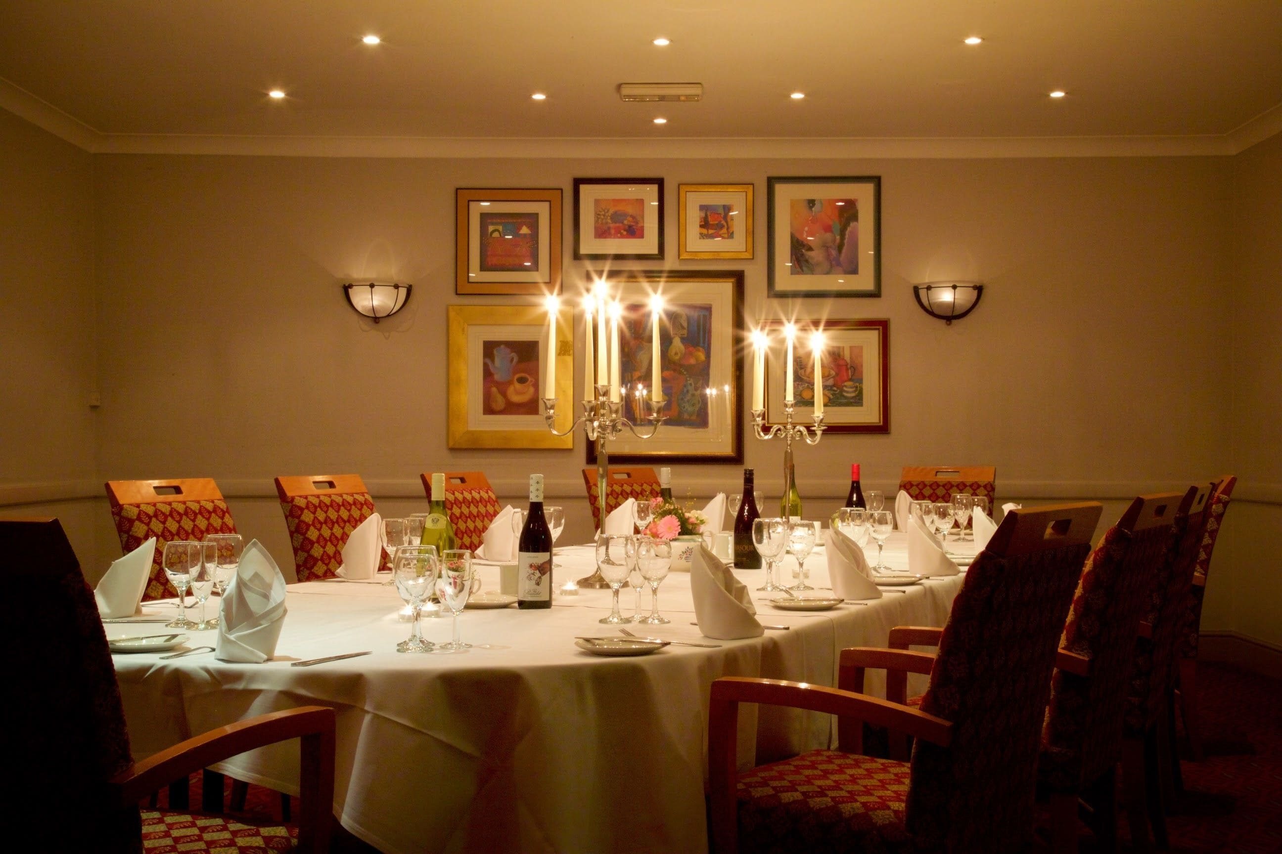A dining room with a long table set for a meal, framed artwork on the walls, and wall-mounted lighting.