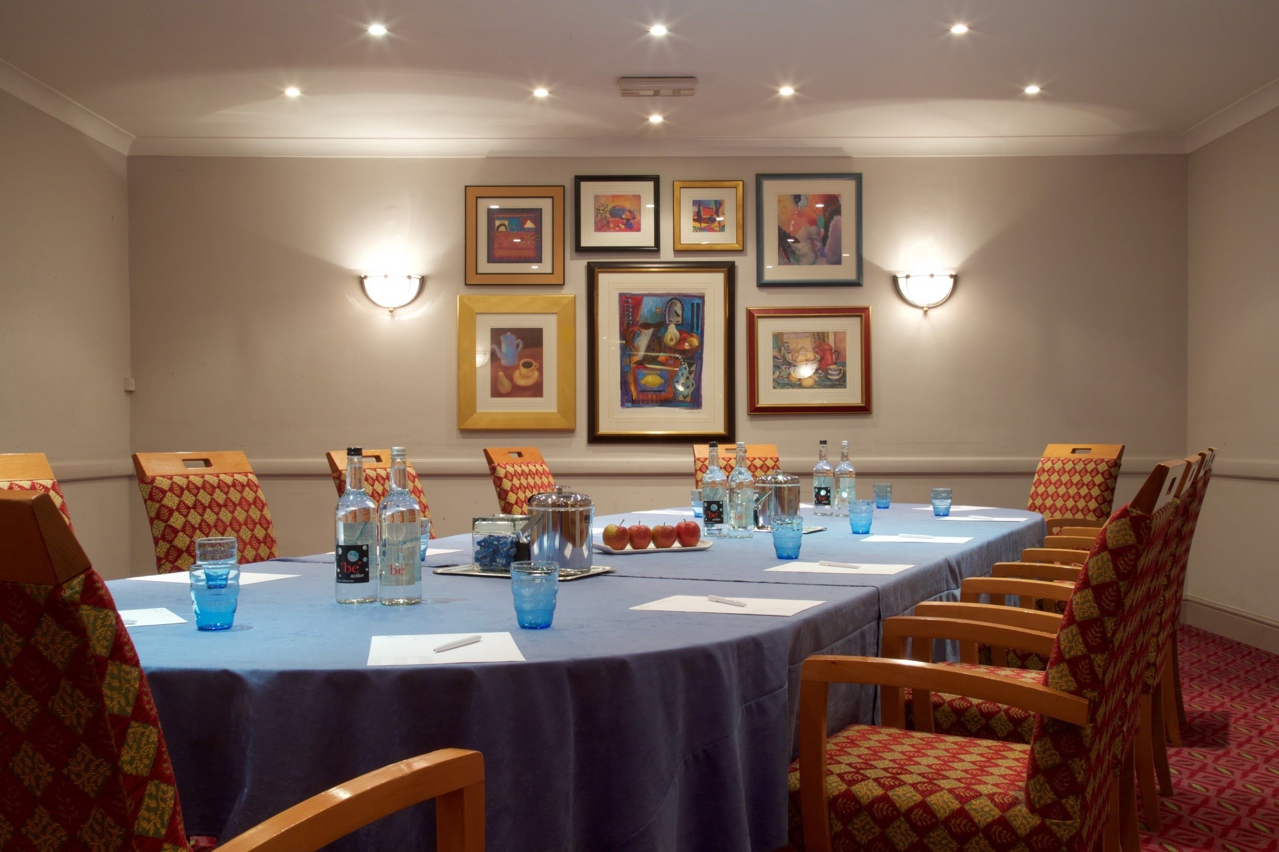 Un salón de conferencias con una larga mesa, sillas, obras de arte enmarcadas en la pared y una disposición de mesa que incluye botellas de agua y vasos.