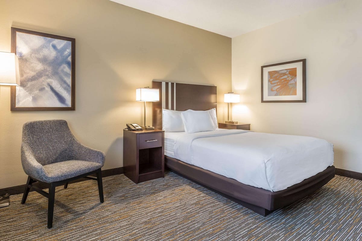 Quarto de hotel com uma cama grande, uma cadeira, mesa de cabeceira com telefone e duas luminárias fixadas na parede.