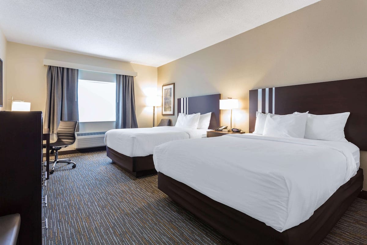 Quarto de hotel com duas camas, uma escrivaninha, uma cadeira e uma janela grande com cortinas.