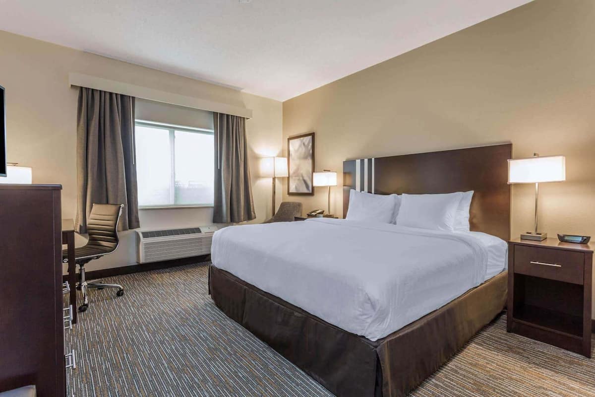 Quarto de hotel com uma cama grande, uma mesa com cadeira, luminária de mesa, telefone e janela com cortinas.