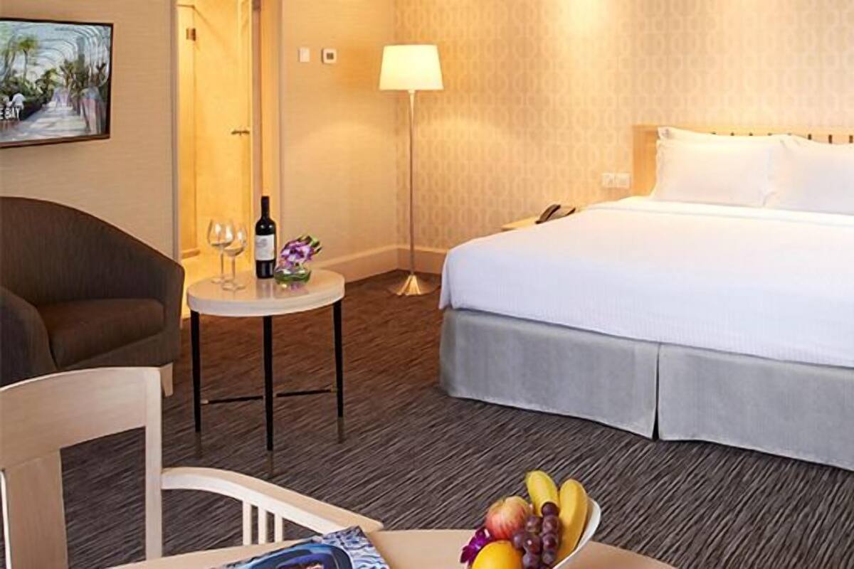 A hotel room with a bed, a chair, a small table with a bottle and glasses, and a fruit bowl.
