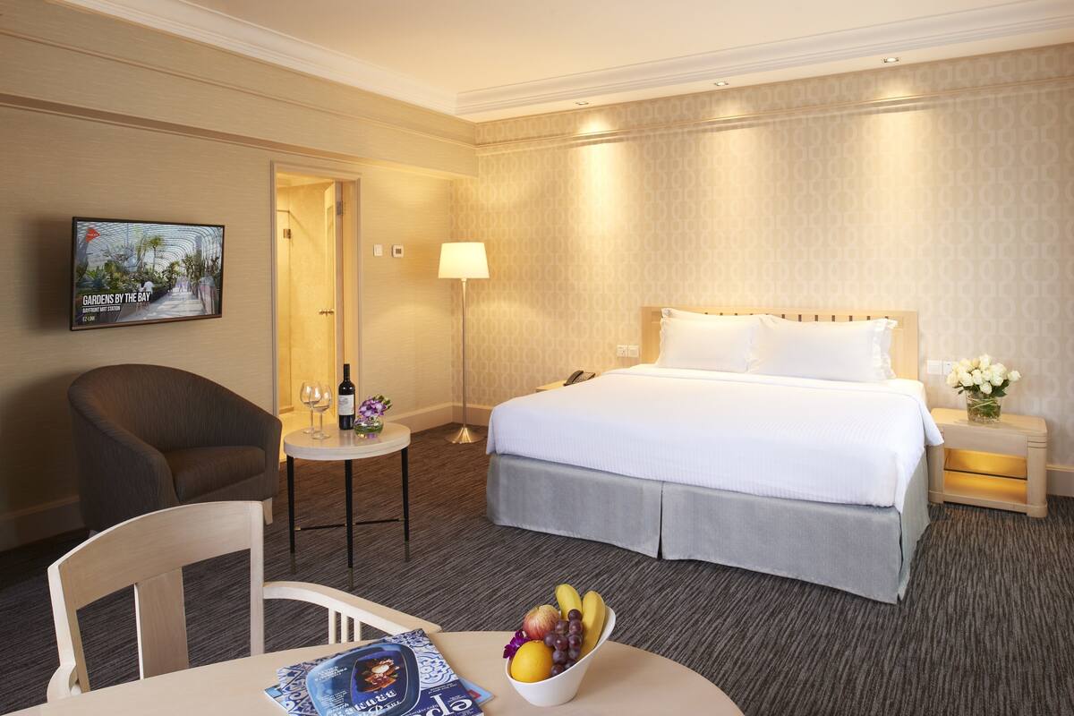 A hotel room with a bed, a desk with a chair, a small table with a bowl of fruit, a lamp, and a TV on the wall.