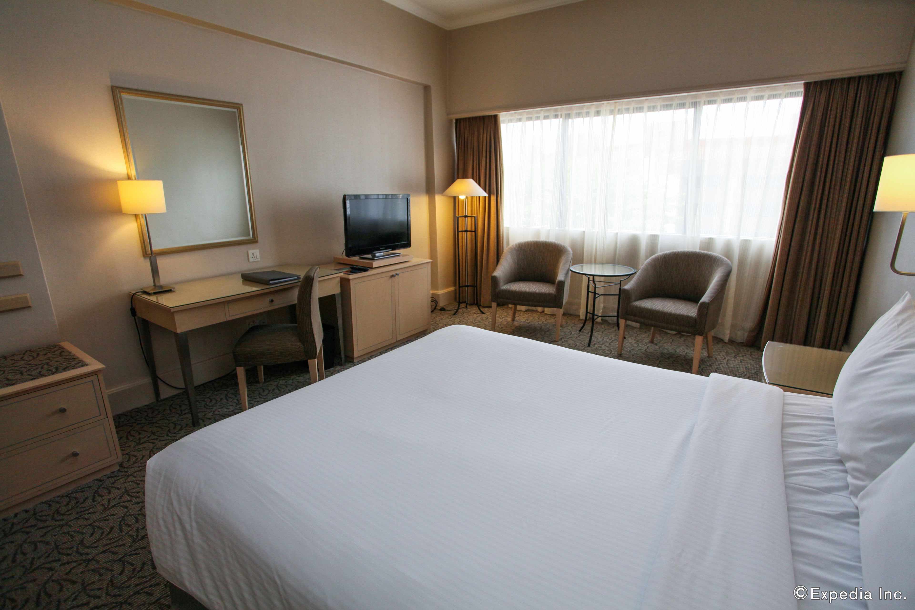 A hotel room with a large bed, a desk with a computer, a TV, two armchairs, and a window with curtains.