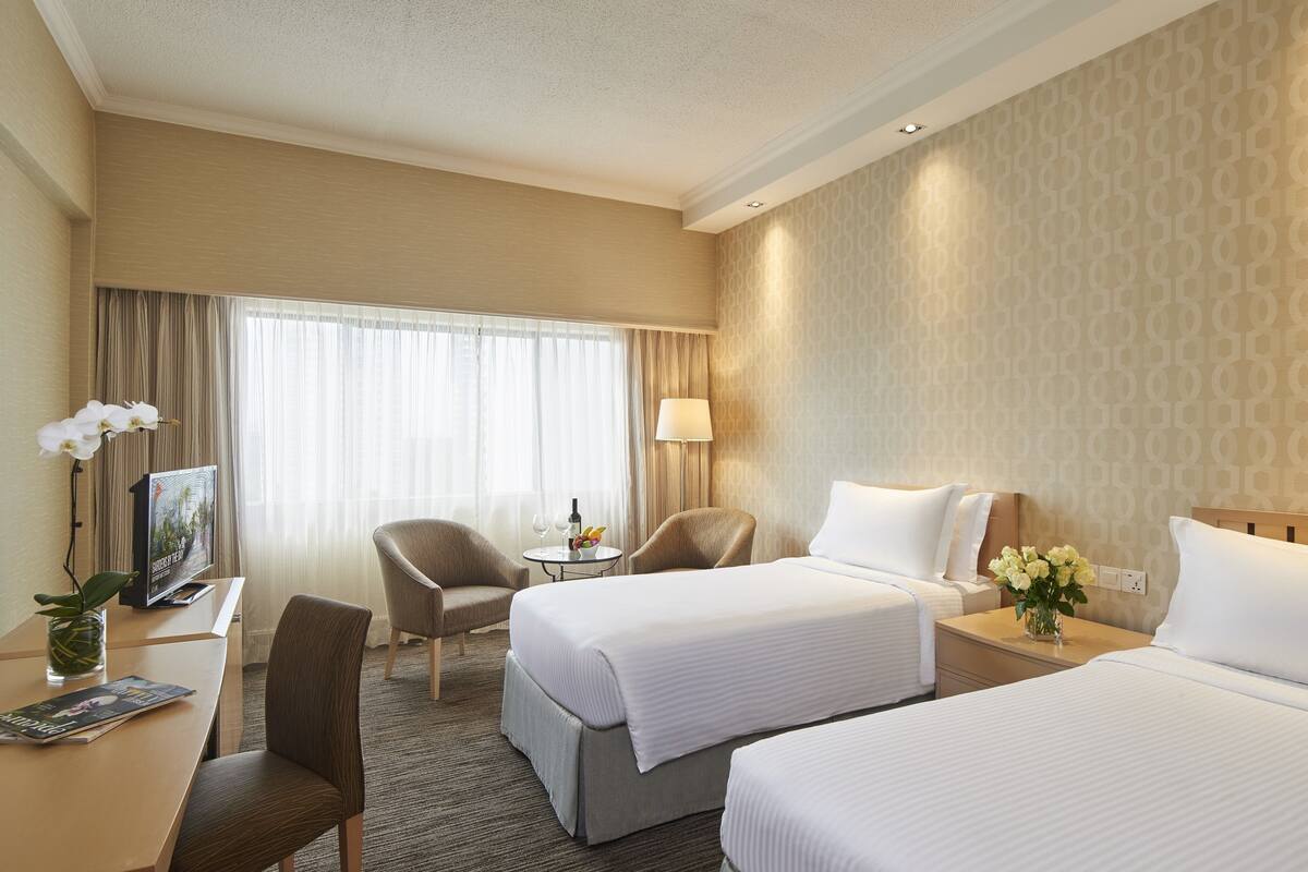 A hotel room with two beds, a desk with a computer and a lamp, a chair, a small table with a vase of flowers, and a window with curtains.