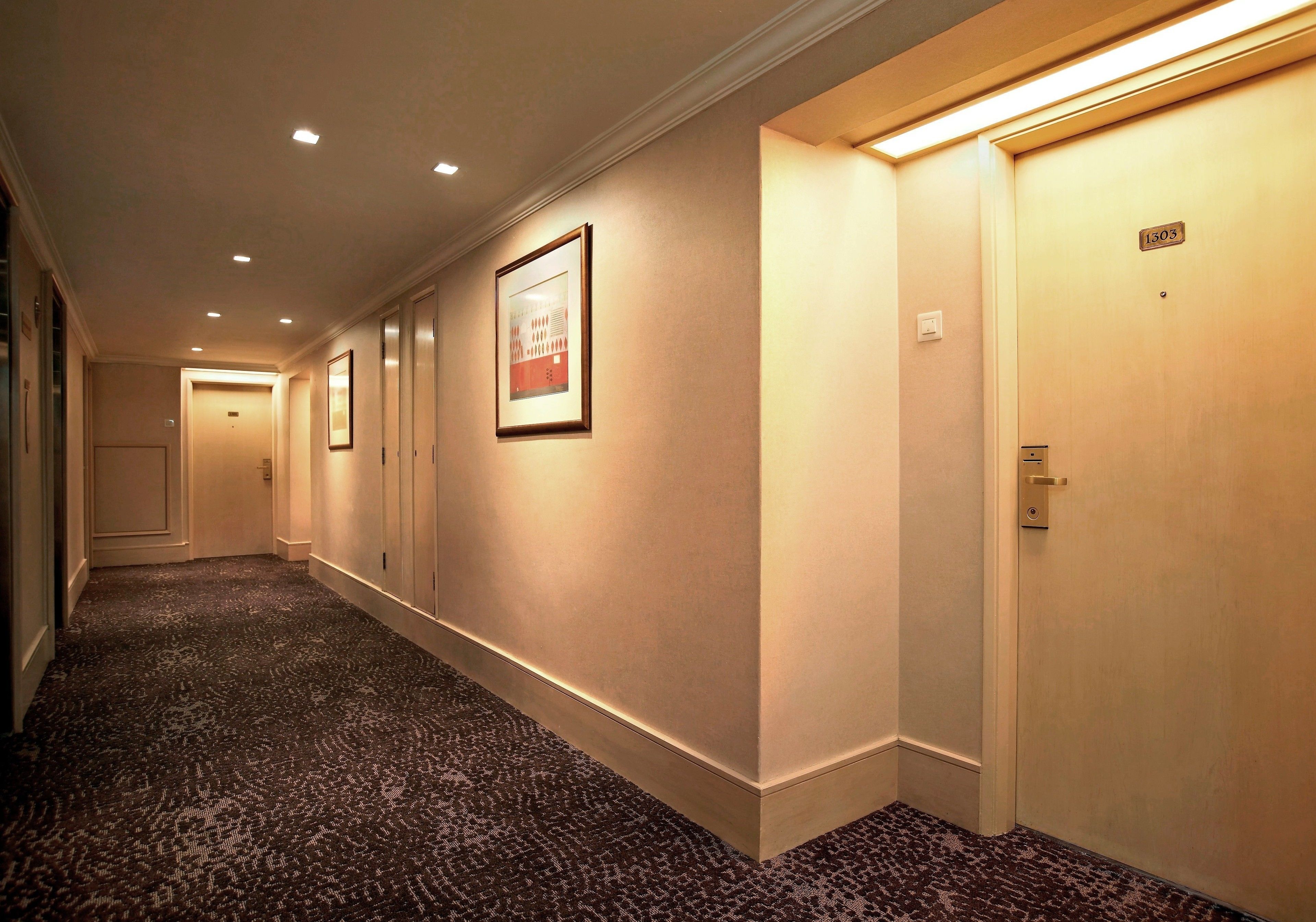 Un pasillo de hotel con pisos alfombrados, cuadros enmarcados en las paredes y puertas numeradas 1303.