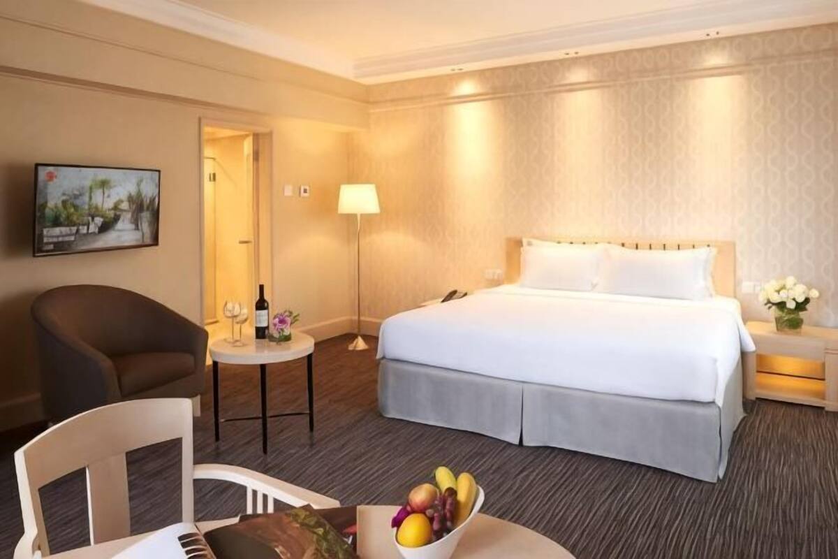 A hotel room with a bed, a chair, a small table with a fruit bowl, a lamp, and a painting on the wall.