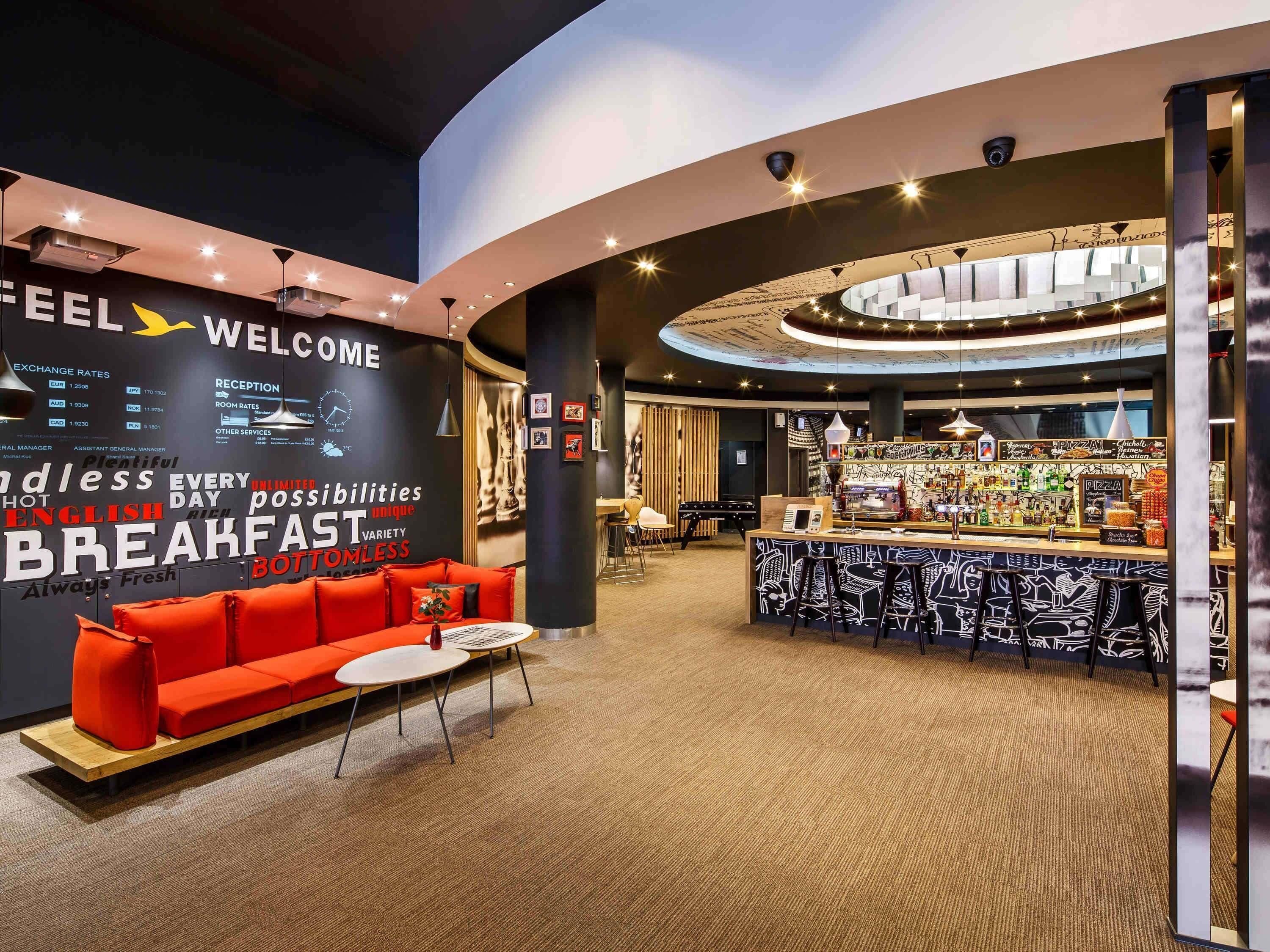 A modern hotel lobby with a curved reception desk, a seating area with red sofas, and a bar with a pizza display.