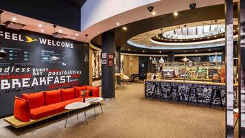 A modern hotel lobby with a curved reception desk, a seating area with red sofas, and a bar with a pizza display.