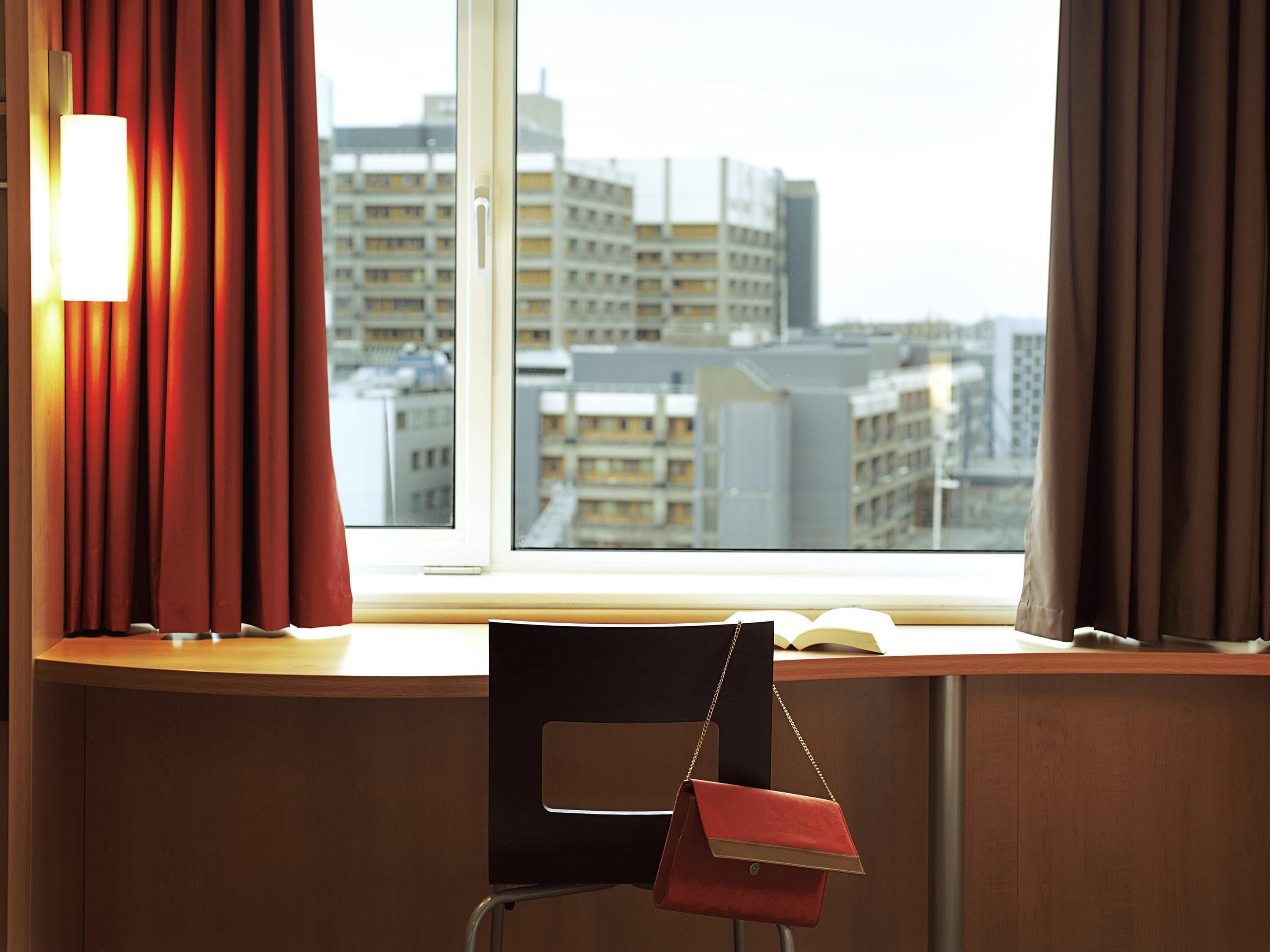 A hotel room with a view of city buildings, a desk with a chair, and a window with curtains.