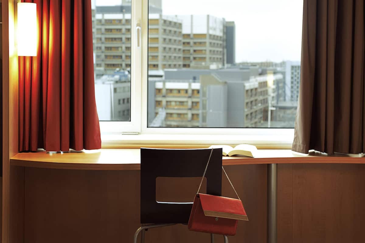 A hotel room with a view of city buildings, a desk with a chair, and a window with curtains.