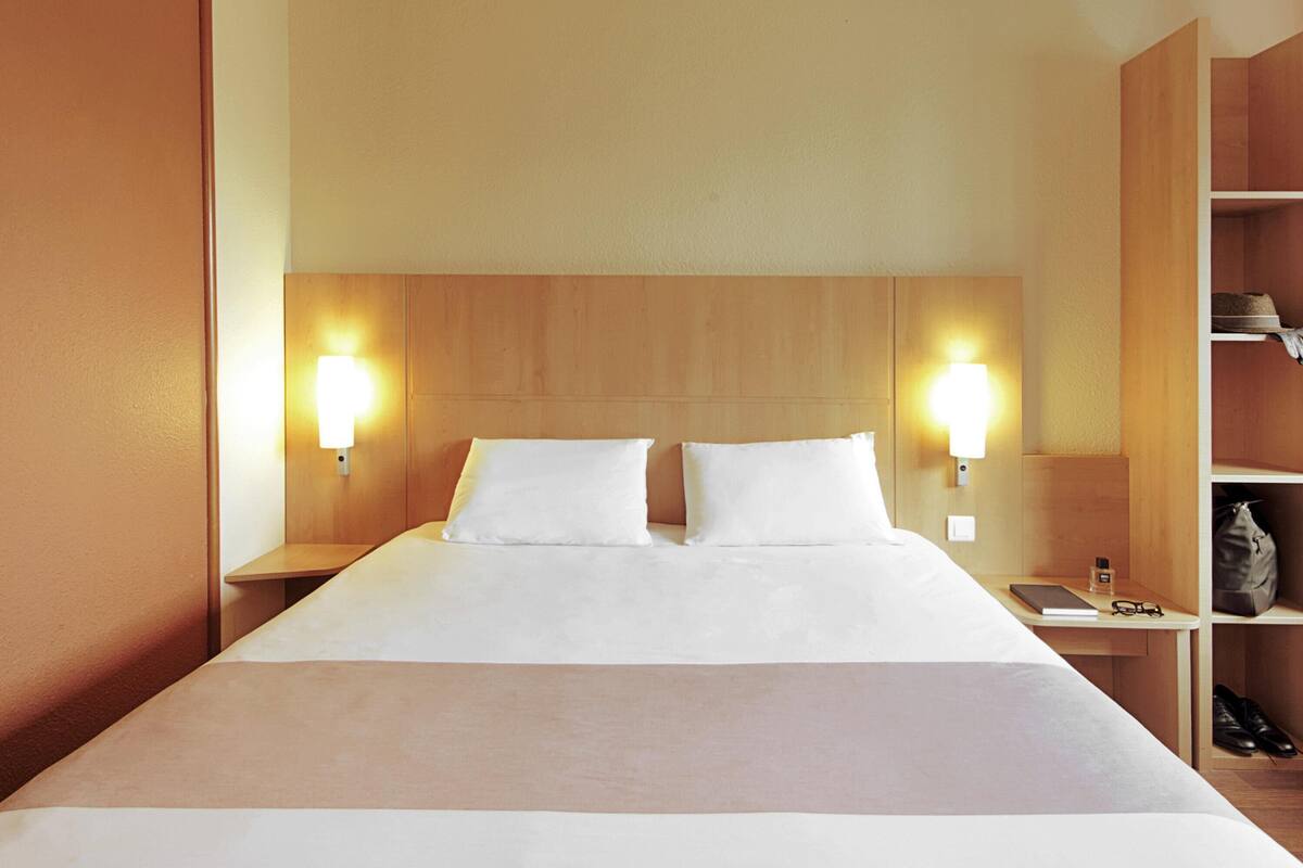 Ein Hotelzimmer mit einem großen Bett, zwei Nachttischlampen, einem Nachttisch und einem Holzregal mit Hut und Tasche.