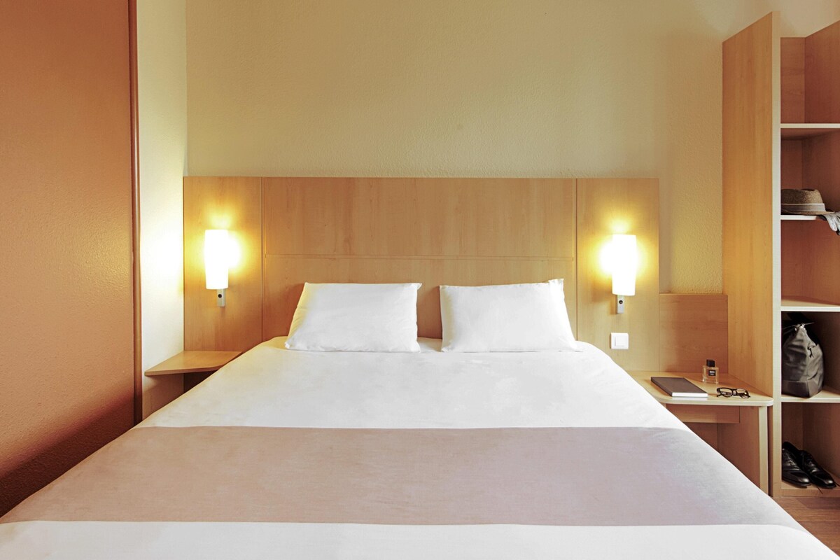 A hotel room with a large bed, two bedside lamps, a nightstand, and a wooden shelf with a hat and a bag.