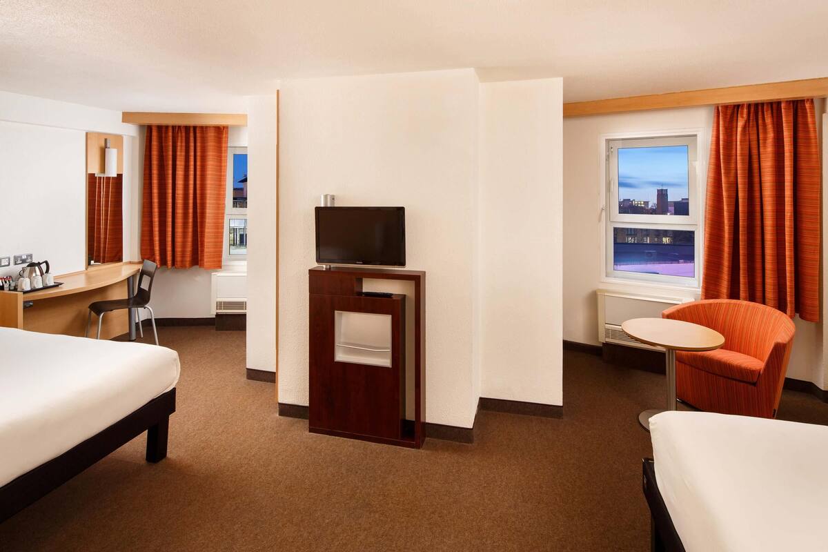 Ein Hotelzimmer mit zwei Betten, einem Schreibtisch, einem Stuhl, einem Fernseher und einem Fenster mit Vorhängen.