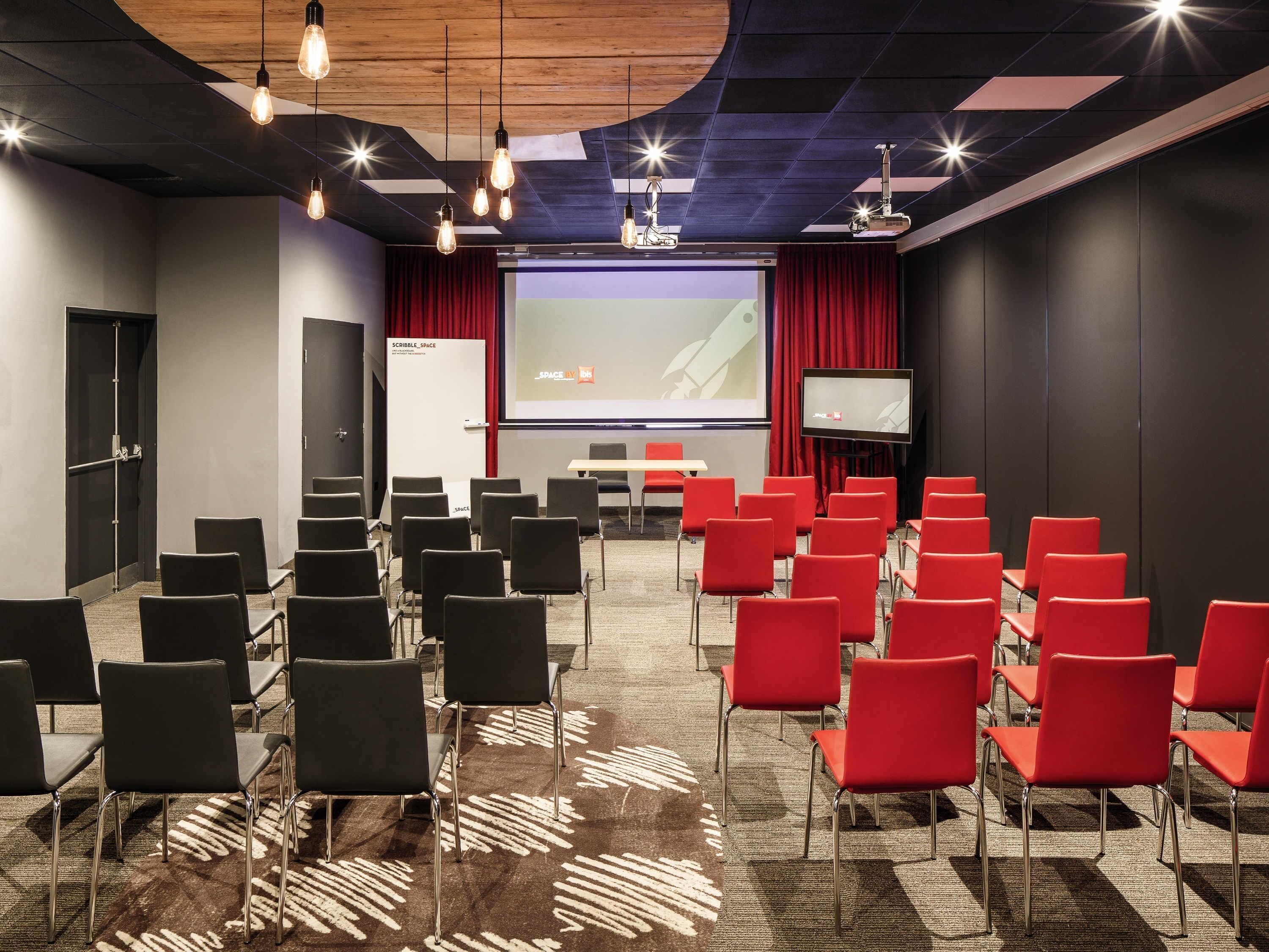 A conference room with rows of chairs, a projector screen, and a screen displaying a presentation.