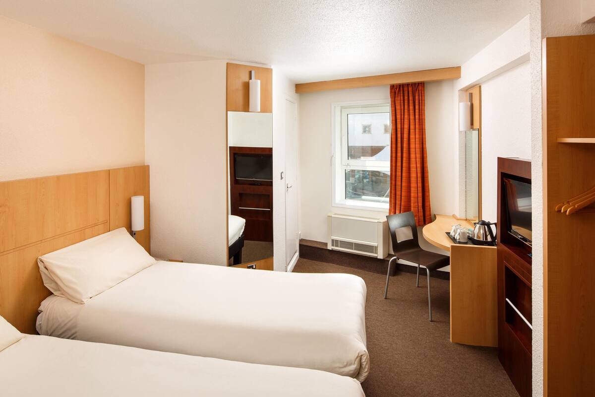 Ein Hotelzimmer mit zwei Betten, einem Schreibtisch mit Stuhl, einem Fernseher und einem Fenster mit Vorhängen.