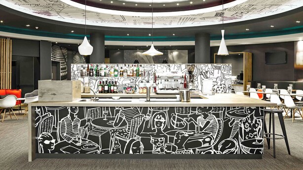 A modern bar area with a circular ceiling design, a bar counter with a black and white mural, and multiple hanging pendant lights. - Ibis Birmingham New Street Station Hotel (Birmingham)
