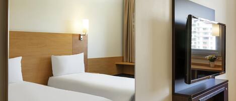A hotel room with two beds, a wooden headboard, a TV mounted on a wooden cabinet, and a window with a view of buildings.